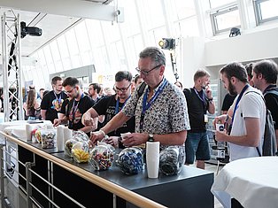 Man in patterned shirt demonstrates LEGO robots to attendees at a bright indoor event with professional lighting and large windows.