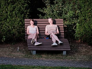 Two women sitting on an oversized wooden bench in a garden at night, surrounded by green foliage.