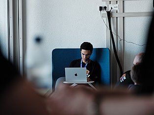 Professional man wearing glasses and lanyard sits on blue chair working on laptop in modern office setting
