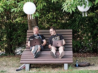 Two men sitting on a bench with laptops in a garden surrounded by green foliage and white spherical lights