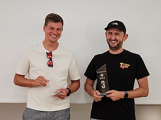 Two smiling men holding awards, one in cream shirt with sunglasses, one in black shirt and cap