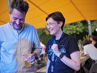 Two people smiling under a yellow tent at an outdoor event, one holding a drink and wearing a lanyard