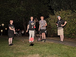 Five people standing on grass at night during an outdoor event with trees and lighting in background