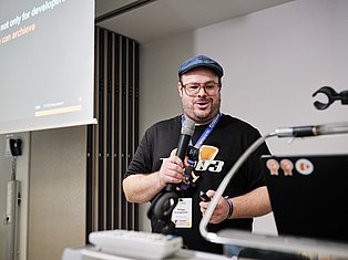 Man with glasses and blue cap speaking at microphone during tech conference presentation