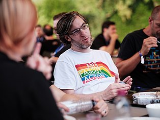 Man wearing anti-fascist rainbow shirt speaking at outdoor community gathering with people around table