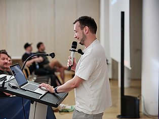 Man in white shirt speaking at podium with microphone and laptop during presentation to seated audience
