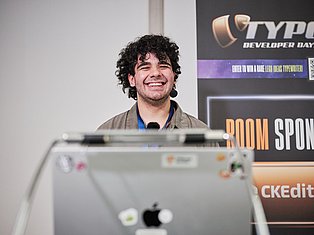 Smiling man with curly hair speaking at a tech developer conference with branded backdrop