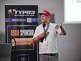 Man speaking at TYPO3 Developer Days 2026 conference with microphone and red cap