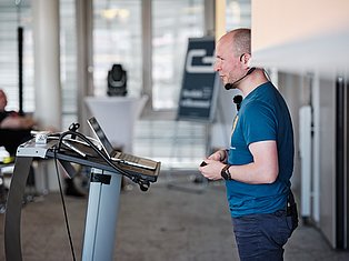 Man in blue shirt wearing headset speaking at podium with microphone in modern office with city view
