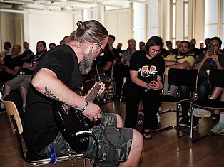 Bearded musician with long hair playing electric guitar while seated, performing for an audience in a bright indoor venue.