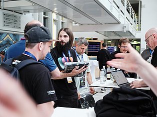 People wearing conference badges gathered around a table discussing technology at an indoor event or conference.