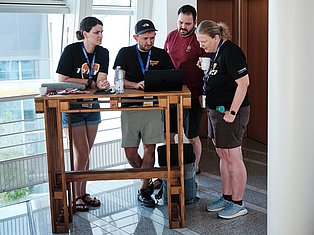 Five people gathered around a wooden table with a laptop, wearing lanyards, collaborating in a bright office with large windows.