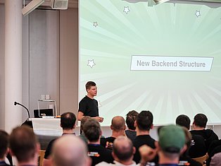 Speaker presenting 'New Backend Structure' to audience at tech conference with projection screen