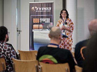 Woman speaking at TYPO3 Developer Days 2020 conference podium with room sponsor banner