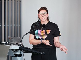 Woman with glasses speaking at podium with microphone and laptop, wearing colorful graphic t-shirt