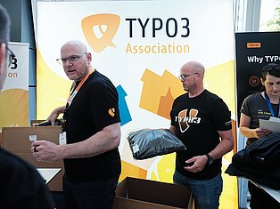 TYPO3 Association representatives presenting at conference booth with yellow branding and merchandise
