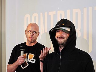 Two men at an event, one wearing glasses holding a microphone, the other in a hoodie and cap, standing before a projected backdrop.