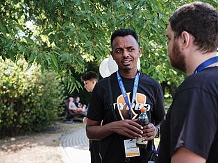 Man wearing conference badge and lanyard talking with colleague outdoors under green trees