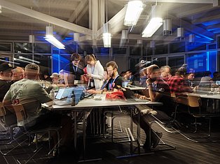 Group of people working at tables in a modern office space with blue lighting and large windows at night