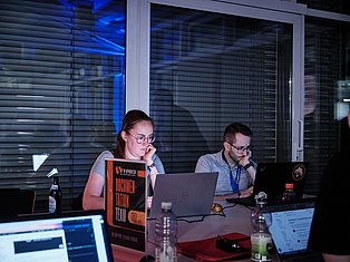 Two developers working at laptops in a modern office with blue lighting and large windows with blinds