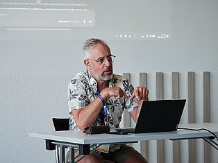 Man with glasses presenting at laptop with projection screen behind him