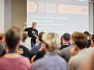 Speaker presenting at conference about room locations, addressing audience with projection screen showing event details