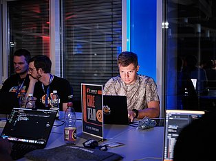 Tech professionals at a conference panel with laptops, wearing ID badges, seated at a table with blue ambient lighting