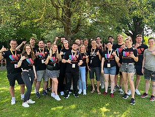 Group of young people wearing matching black t-shirts with the TYPO3 logo and lanyards posing together under a large tree on grass