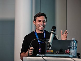 Man speaking at conference with microphone and lanyard badge, smiling and gesturing expressively