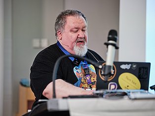 Bearded man speaking at microphone wearing headset and graphic t-shirt at conference table