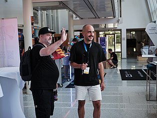 Two men at an indoor event venue, one wearing a Yankees cap waving, the other wearing a badge and black shirt smiling