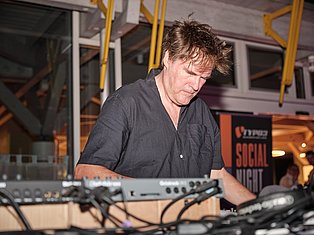 DJ performing at a social night event, operating audio equipment and mixer at a public venue with yellow handrails visible.