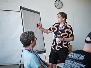 Person in colorful patterned shirt presenting at whiteboard to audience members wearing lanyards in office setting