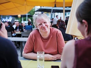 Smiling woman in pink shirt conversing at outdoor event under yellow umbrella with crowd in background