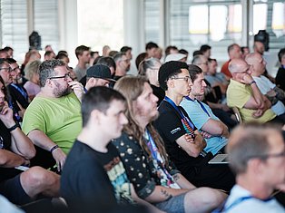 Large diverse crowd of attendees wearing lanyards at an indoor conference or event venue with floor-to-ceiling windows.