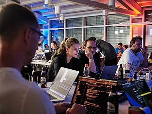 Tech professionals collaborating at a modern office event with laptops, code displays, and colorful ambient lighting in a contemporary workspace.