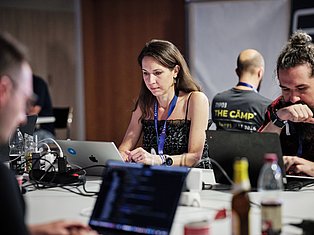 Woman with ID badge working on laptop at conference table with colleagues and tech equipment