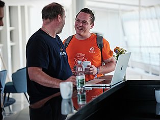 Two men having a friendly conversation at a table with laptops and flowers in a bright office setting.