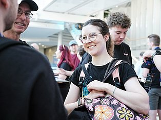 Smiling young woman with glasses at indoor event surrounded by people wearing casual clothing and backpacks