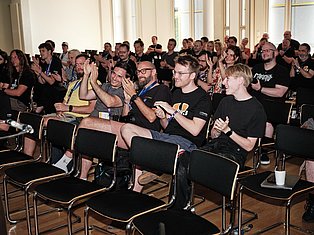 Large audience of people seated and standing in a bright hall, applauding and smiling during a presentation or event.