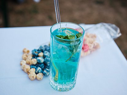Turquoise cocktail with mint leaves and straw, served with berries and snacks on white table