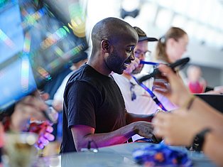 Man in black shirt smiling at social gathering with colorful lights and people holding drinks