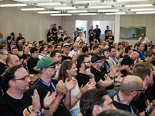 Large crowd of people at indoor conference or event, many wearing lanyards, applauding in modern venue with fluorescent lighting.