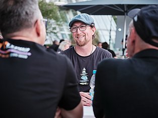 Man wearing blue cap and glasses smiling while conversing with others at an outdoor event under a tent