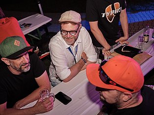 Men at a table during an indoor event, wearing caps and casual clothing, socializing with drinks