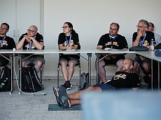 Panel of professionals wearing branded shirts and lanyards seated at a table during a conference or event discussion.