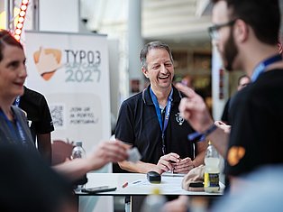 Man with conference badge smiling while talking with attendees at TYPO3 2027 event