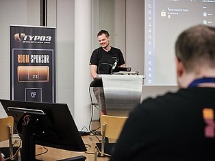 Man speaking at podium during TYPO3 Developer Days 2020 conference with room sponsor banner and audience