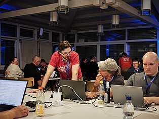 People at a tech conference or hackathon working on laptops at tables with blue ambient lighting and large windows in background.
