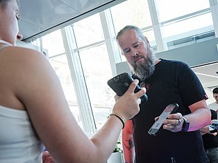 Man with beard holding gaming controllers talking with another person in bright indoor space